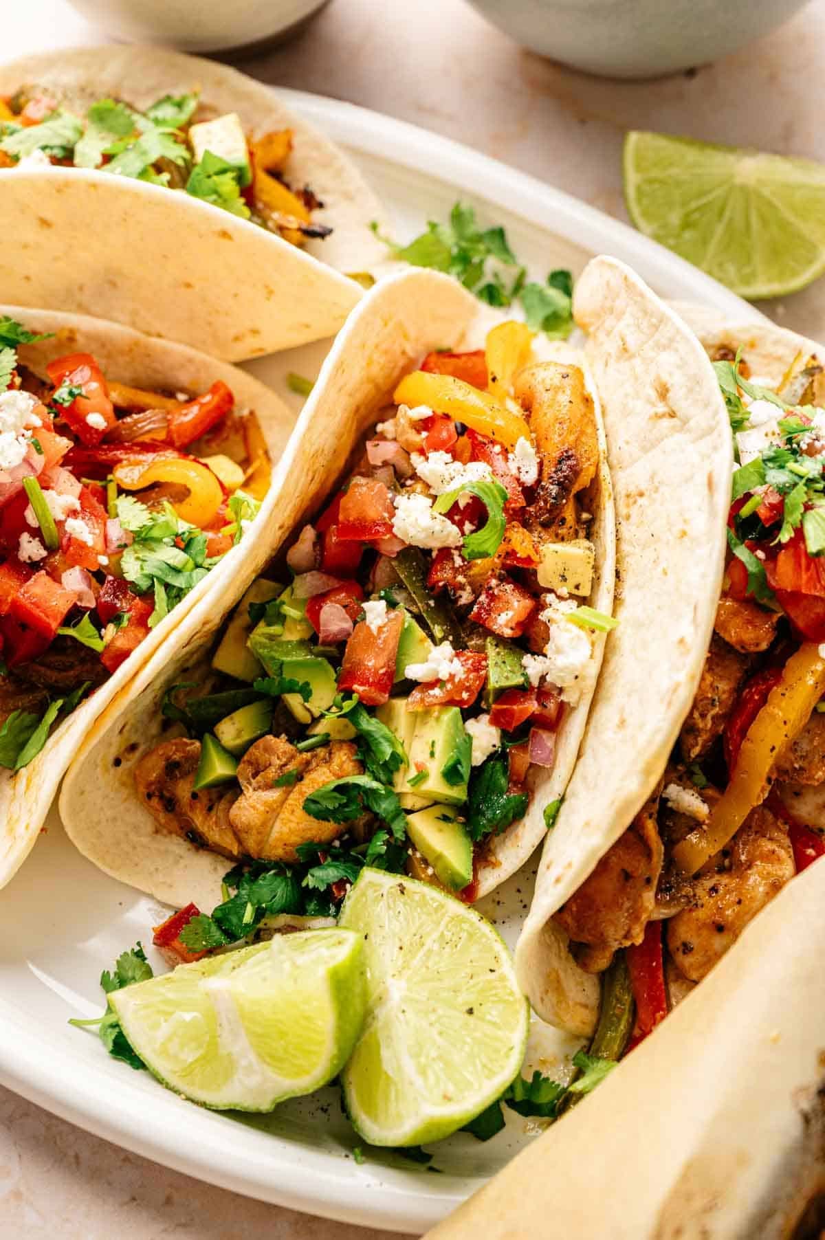 Three soft tacos filled with grilled chicken, diced avocado, tomatoes, onions, cilantro, and crumbled cheese are served on a plate with lime wedges&mdash;perfect for fans of Sheet Pan Fajitas.