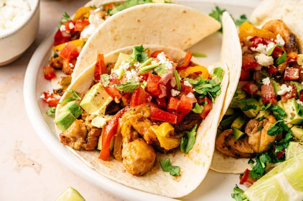 Three chicken Sheet Pan Fajitas tacos topped with diced avocado, tomatoes, peppers, cilantro, and crumbled cheese, served on a white plate with lime wedges.