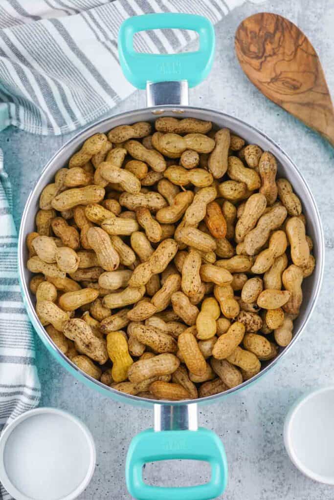 A large pot of Stove Top Boiled Peanuts sits on a countertop, surrounded by a striped towel, a wooden spoon, and two empty white bowls.