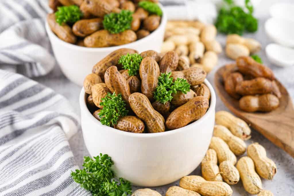 Two white bowls filled with Stove Top Boiled Peanuts, garnished with parsley, sit on a striped cloth with unshelled peanuts scattered around.