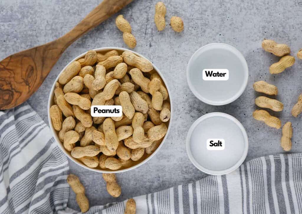 A bowl of unshelled peanuts, a small bowl of salt, and a small bowl of water arranged on a gray surface with a striped cloth and wooden spoon&mdash;perfect ingredients for making classic Stove Top Boiled Peanuts.