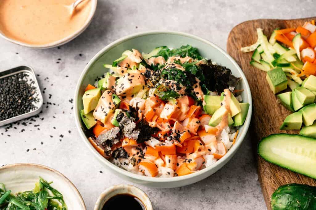 A bowl of poke with diced salmon, avocado, carrot, seaweed, and black sesame seeds on rice, inspired by the flavors of a California Roll Sushi Bowl and surrounded by chopped vegetables and sauce bowls.