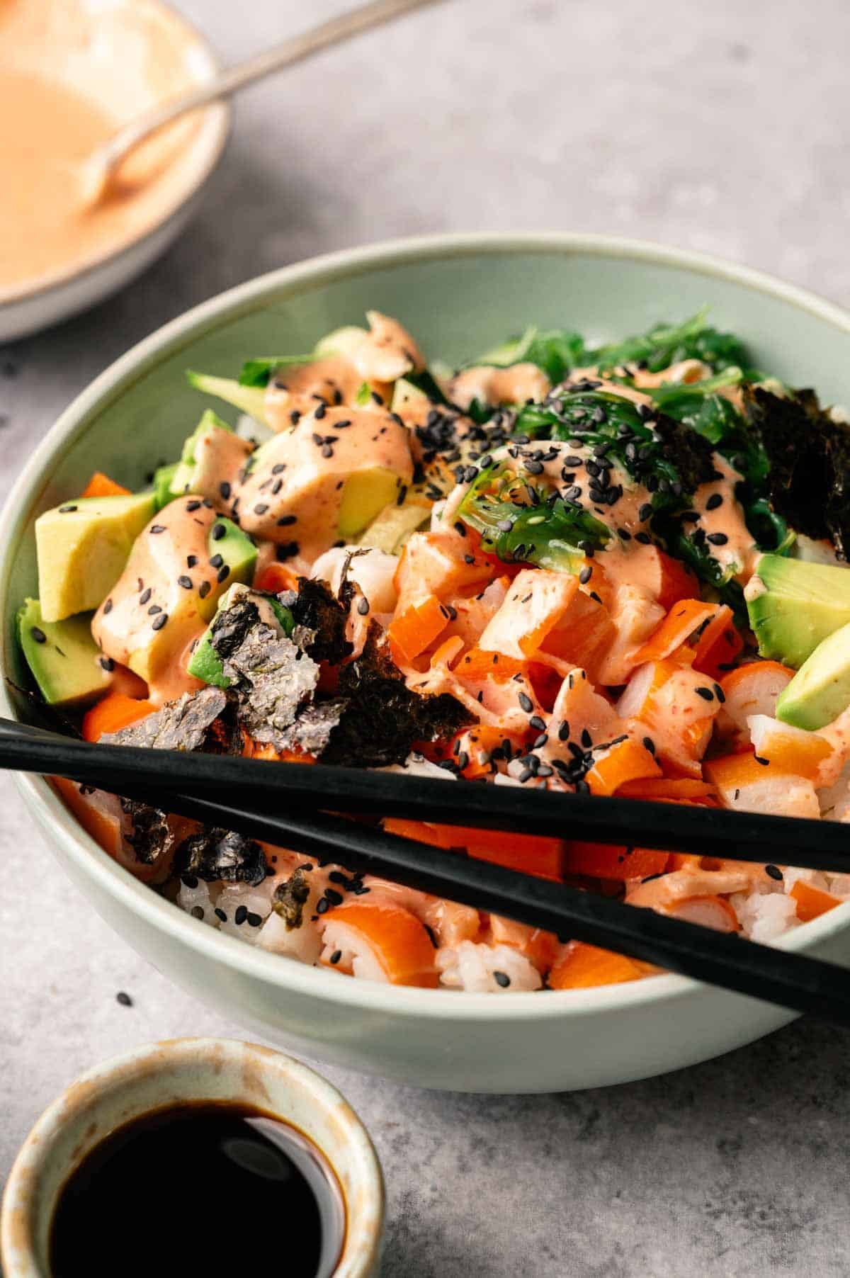 A California Roll Sushi Bowl featuring diced salmon, avocado, seaweed, rice, black sesame seeds, and sauce, served with chopsticks and a small dish of soy sauce.