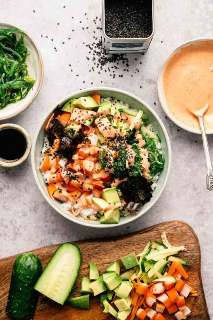 Experience a California Roll Sushi Bowl: a bowl of rice topped with diced vegetables, avocado, seaweed, and sesame seeds, surrounded by sliced cucumber, extra veggies, savory sauce, soy sauce, and a container of black sesame seeds.