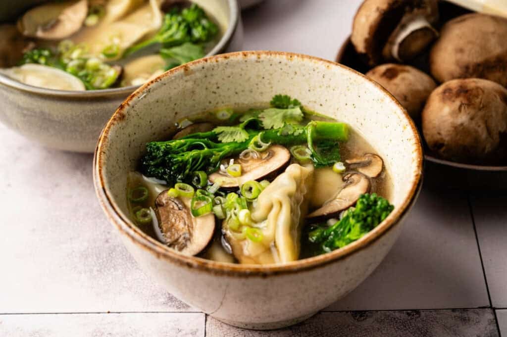 A bowl of wonton soup with dumplings, sliced mushrooms, broccoli, chopped green onions, and fresh cilantro. A small bowl of mushrooms sits beside it.