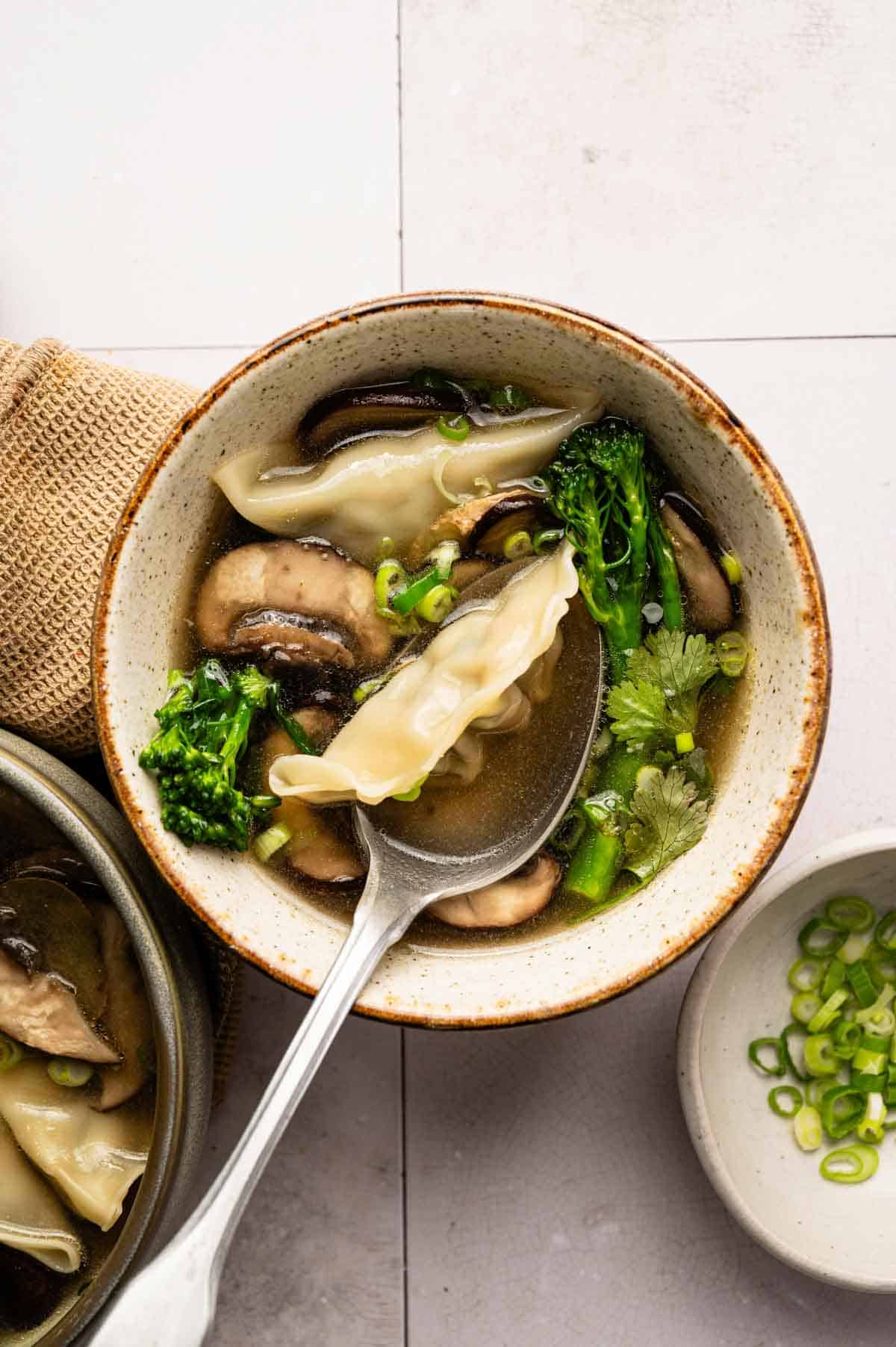 A bowl of Wonton Soup with dumplings, mushrooms, broccolini, fresh herbs, and sliced green onions, with a spoon lifting a dumpling.