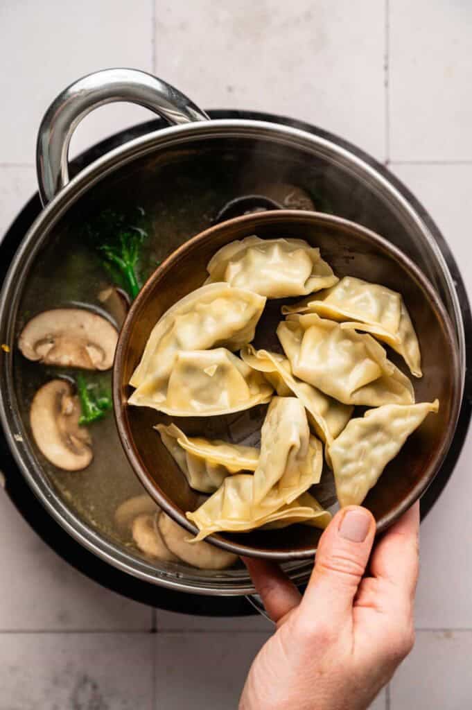 A hand holds a bowl of uncooked dumplings above a pot of wonton soup filled with mushrooms and greens.