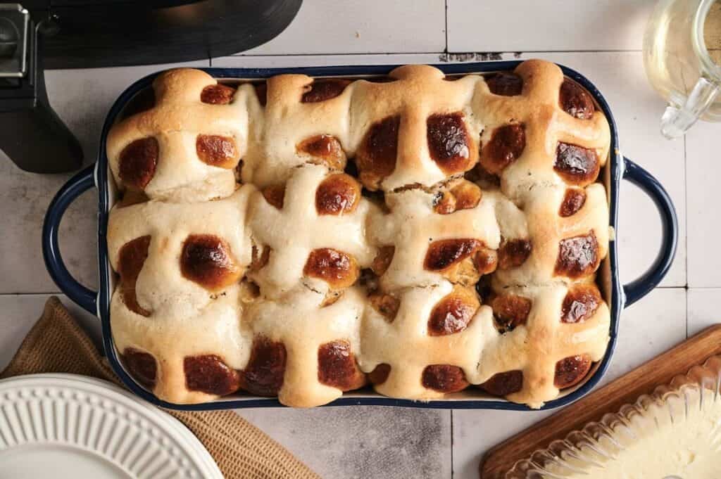 A baking dish filled with golden brown Air Fryer Hot Cross Buns, topped with white icing crosses, sits on a tiled surface next to plates and a glass teapot.