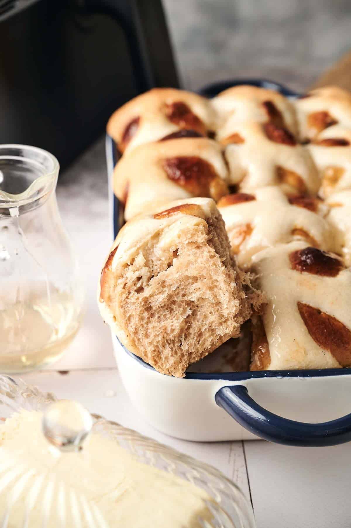 A baking dish filled with glazed Air Fryer Hot Cross Buns, with one bun pulled out to reveal its soft, fluffy texture. A glass jug and butter dish sit nearby.