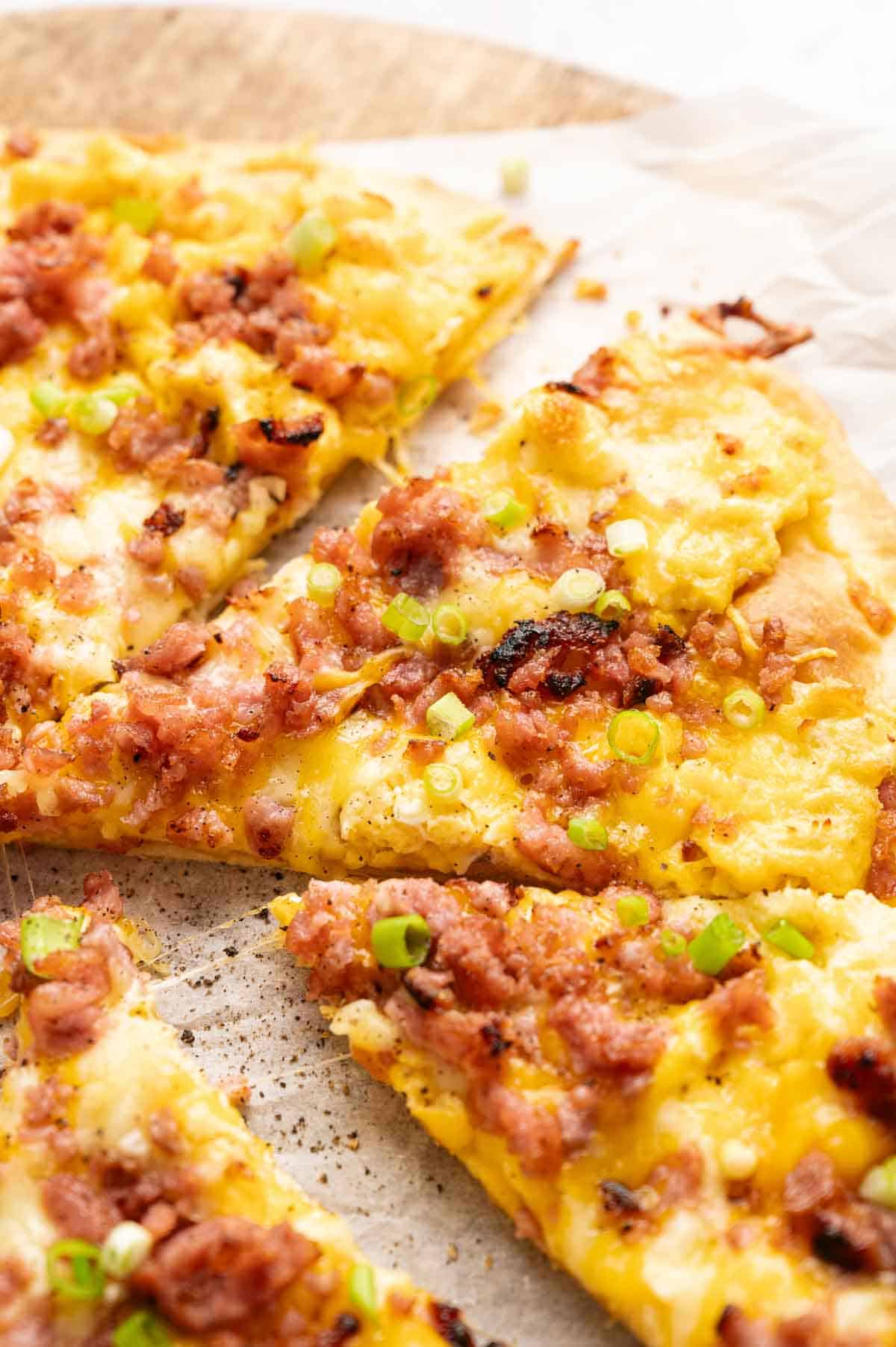 Close-up of Breakfast Pizza slices topped with scrambled eggs, crumbled sausage, melted cheese, and chopped green onions on parchment paper.