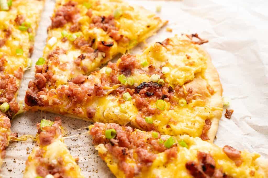 A close-up of sliced Breakfast Pizza topped with scrambled eggs, bacon, cheese, and chopped green onions on parchment paper.
