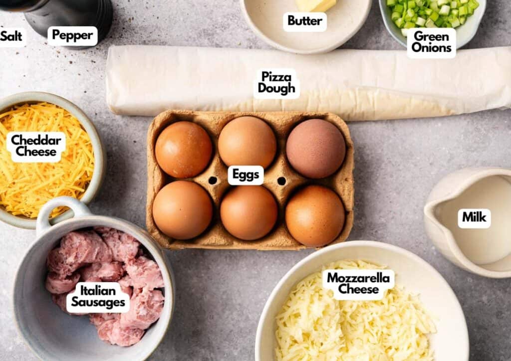 Top-down view of labeled ingredients for a Breakfast Pizza recipe, featuring eggs, pizza dough, butter, green onions, milk, mozzarella cheese, Italian sausages, cheddar cheese, salt, and pepper.