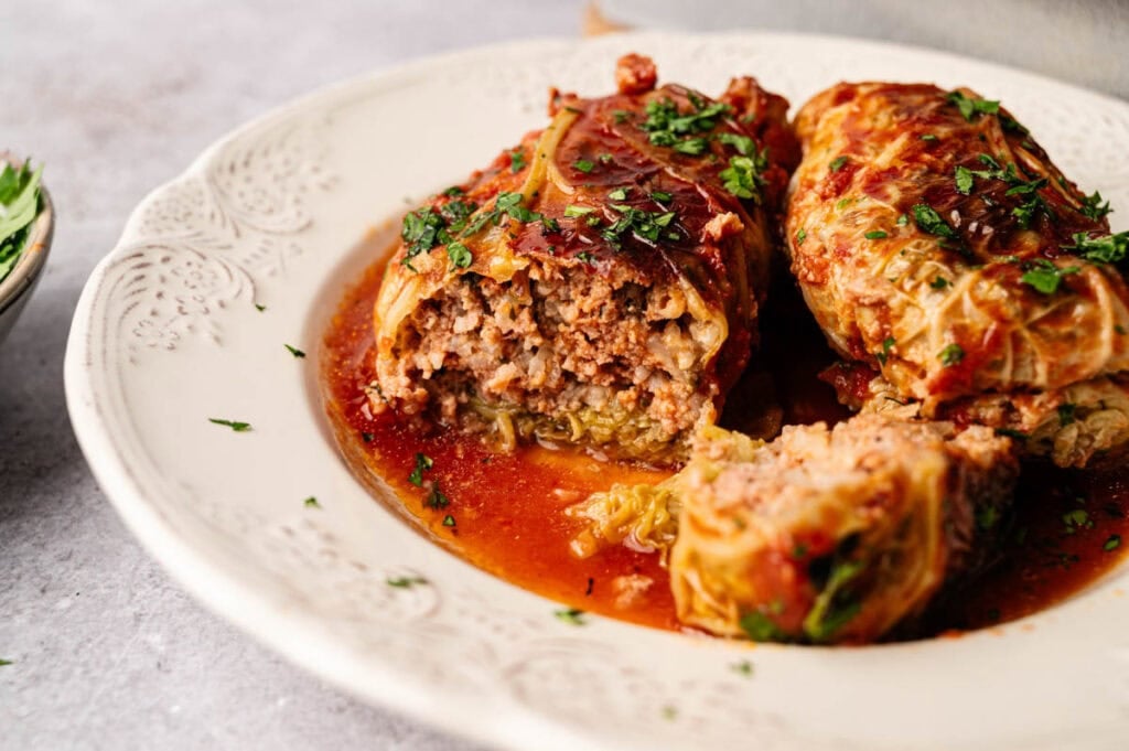 Two Cabbage Rolls covered in tomato sauce, garnished with chopped herbs, served on a decorative white plate. One roll is cut open, revealing the savory meat filling inside.