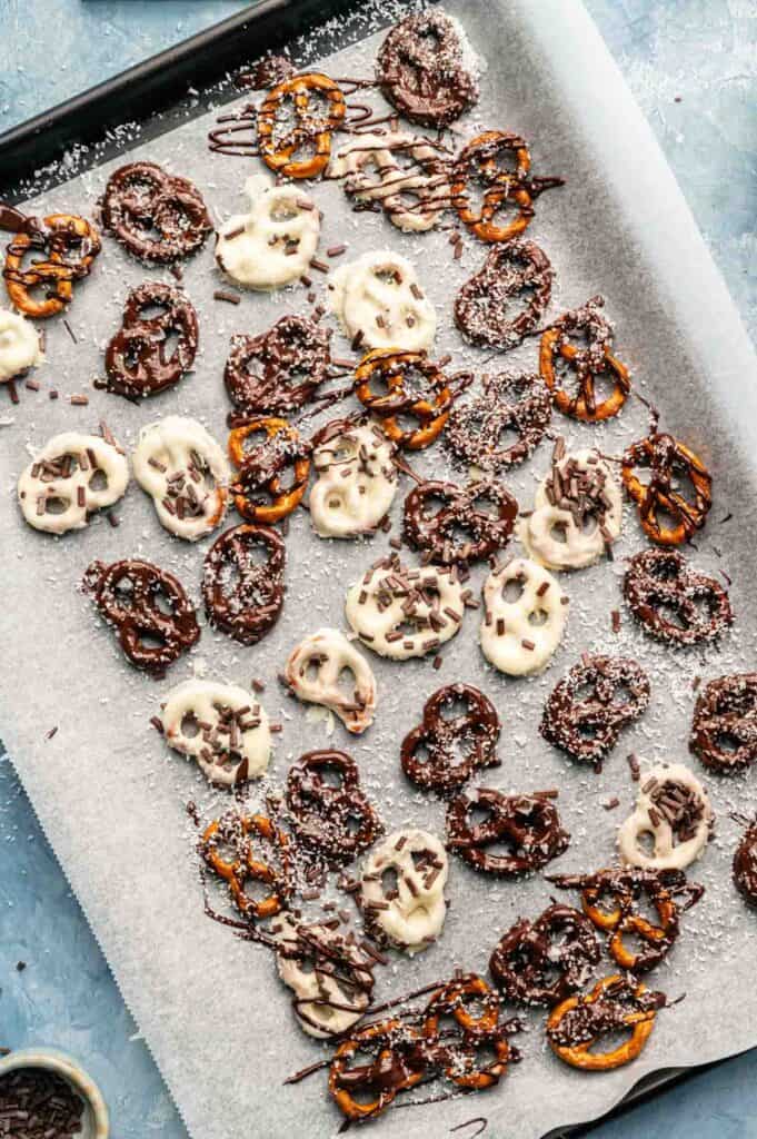 A baking tray lined with parchment paper holds chocolate covered pretzels dipped in white and dark chocolate, then sprinkled with shredded coconut and chocolate sprinkles.