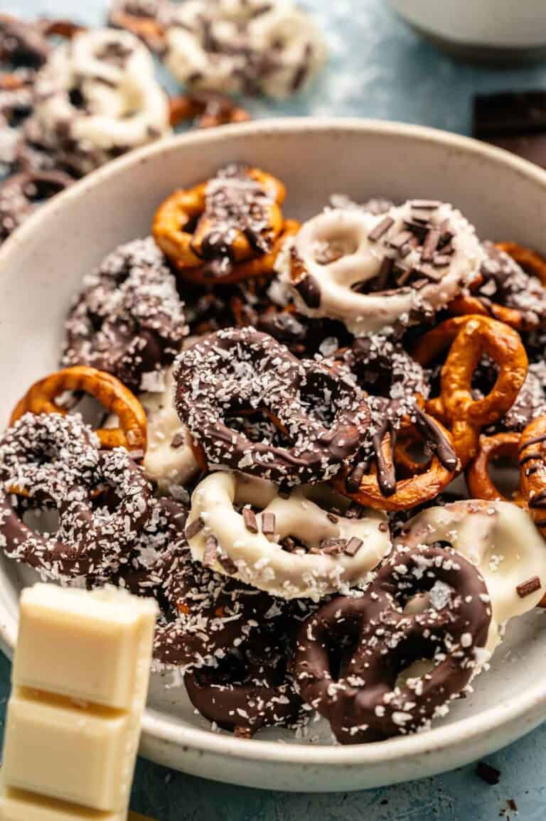 A bowl of Chocolate Covered Pretzels coated in white and dark chocolate, topped with shredded coconut and chocolate sprinkles, with pieces of white chocolate nearby.