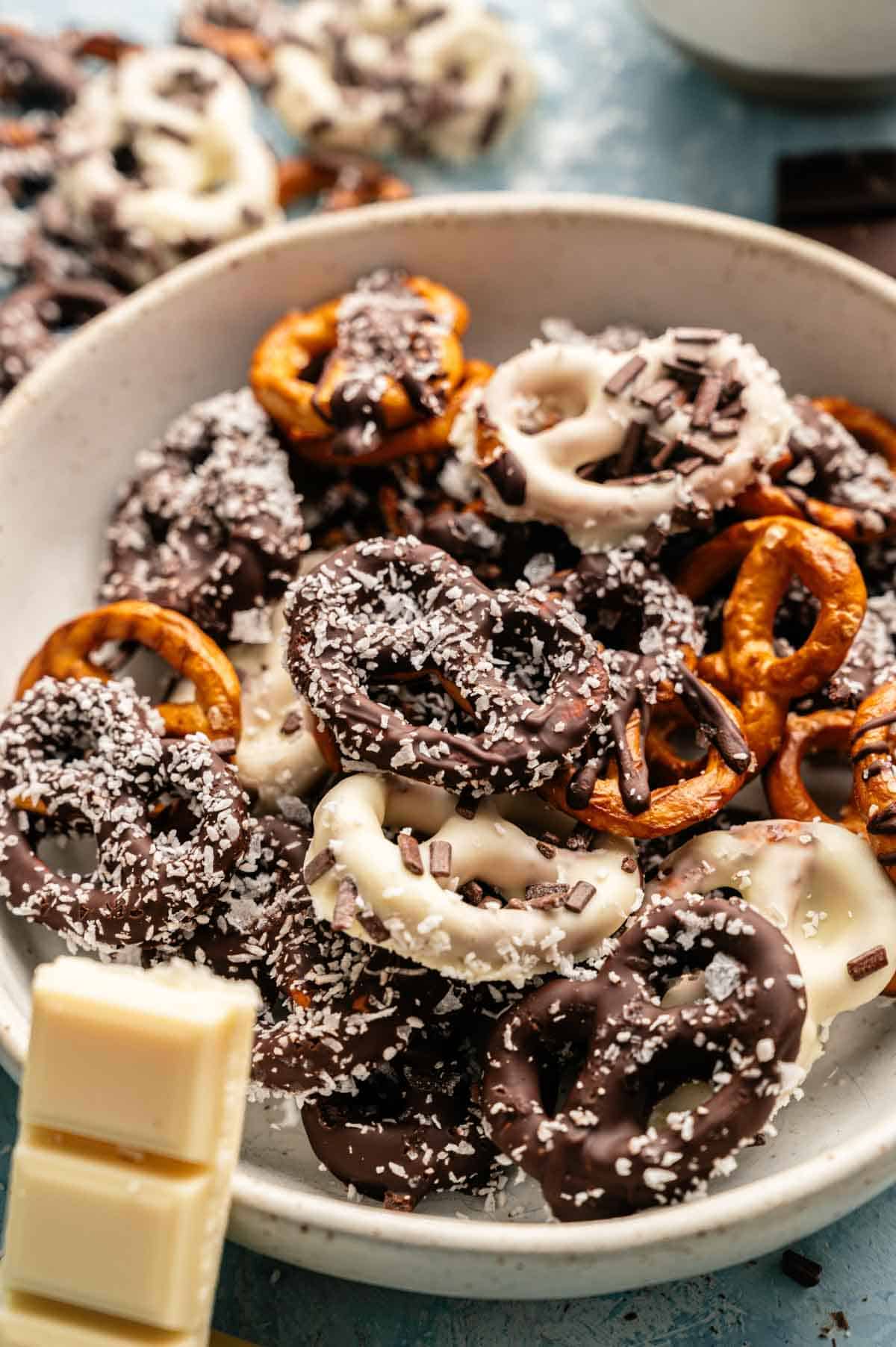 A bowl of Chocolate Covered Pretzels coated in white and dark chocolate, topped with shredded coconut and chocolate sprinkles, with pieces of white chocolate nearby.