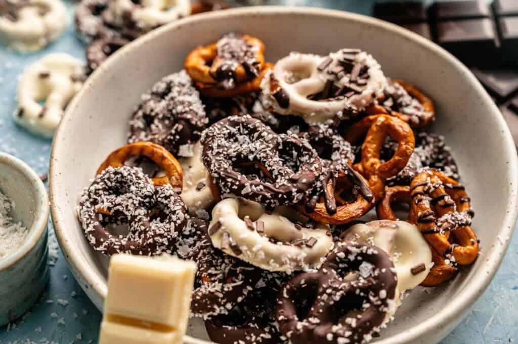 A bowl of Chocolate Covered Pretzels, coated in dark and white chocolate, topped with shredded coconut and chocolate sprinkles, sits on a light blue surface.