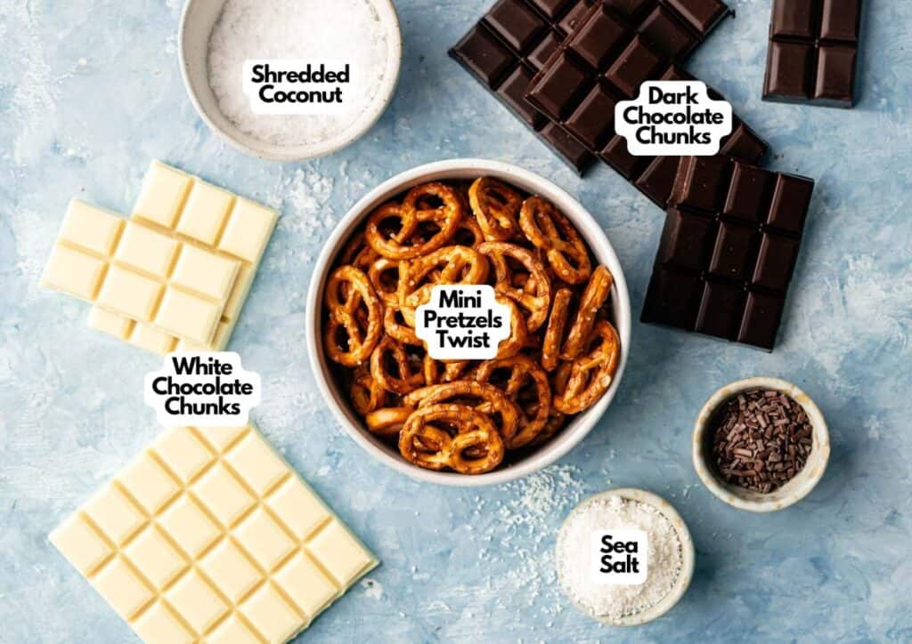 A bowl of mini pretzel twists, perfect for making Chocolate Covered Pretzels, surrounded by white and dark chocolate chunks, shredded coconut, sea salt, and a bowl of chocolate sprinkles on a blue surface.