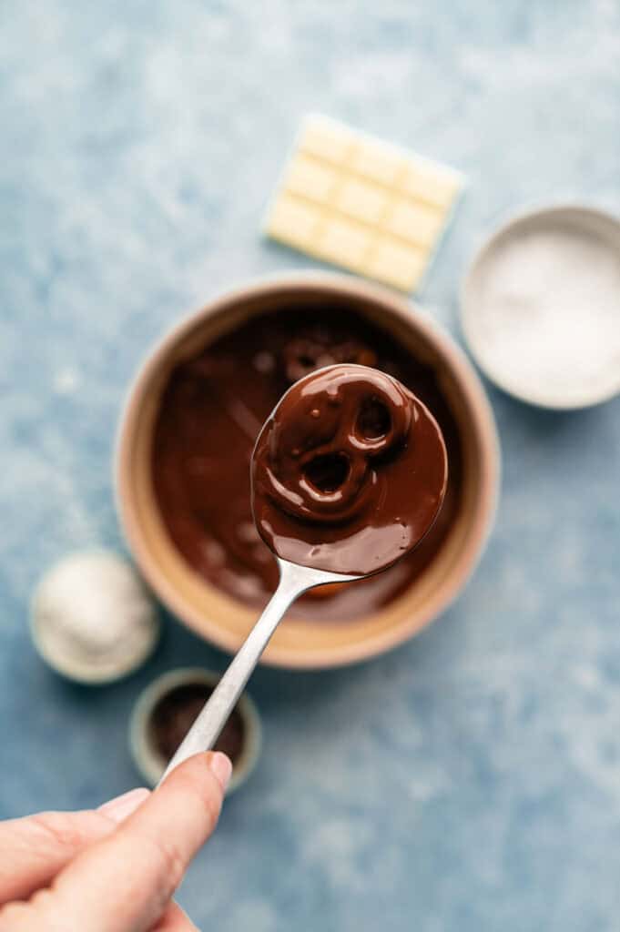 A hand holds a spoon with melted chocolate above a bowl, ready to coat chocolate covered pretzels, with white chocolate and bowls of ingredients in the background.