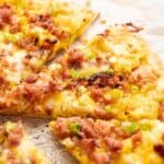 A close-up of a Breakfast Pizza topped with cheese, crumbled sausage, and chopped green onions on parchment paper.