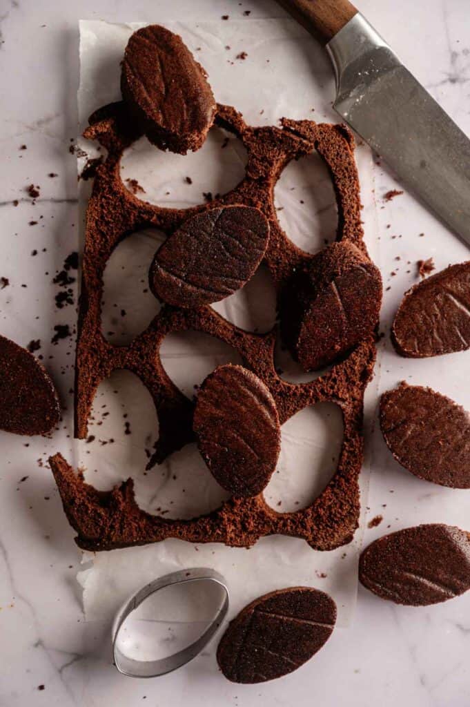 Football Shaped Brownies are cut from a sheet of chocolate dough on parchment paper, with a metal cookie cutter and knife nearby.