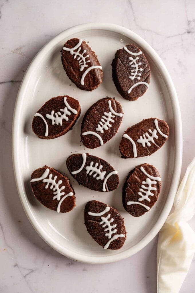 An oval white plate holds eight Football Shaped Brownies, each decorated with white icing to resemble football laces.