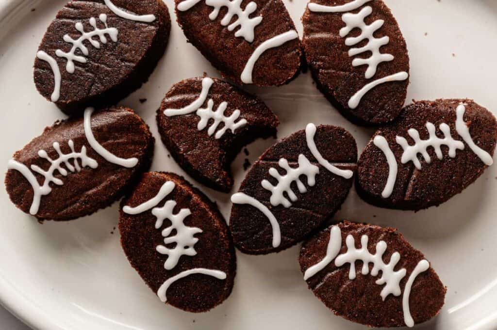 Football Shaped Brownies decorated with white icing to resemble footballs, arranged on a white plate. One brownie has a bite taken out of it.