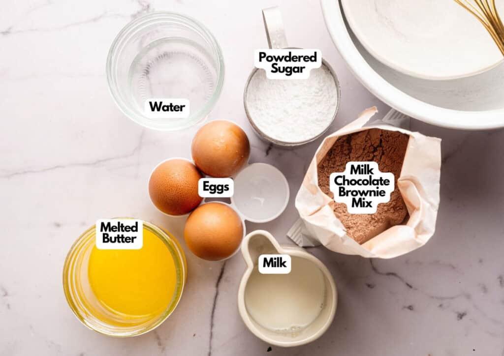 Top-down view of baking ingredients on a marble surface, each labeled for making Football Shaped Brownies&mdash;featuring eggs, water, melted butter, milk, powdered sugar, and milk chocolate brownie mix.