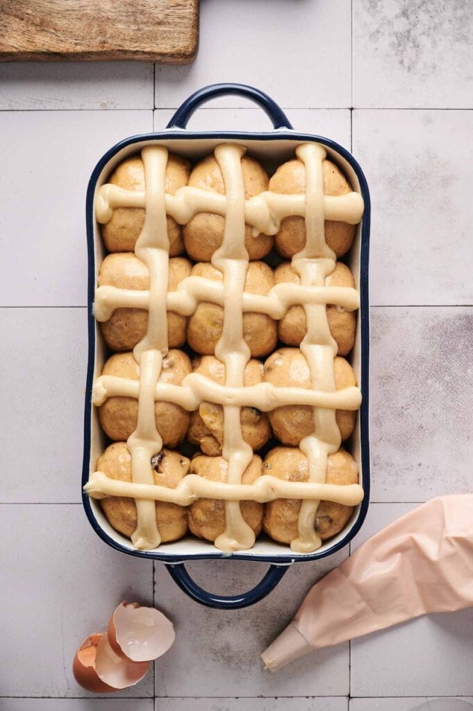 A baking dish filled with uncooked Air Fryer Hot Cross Buns, topped with piped dough crosses. Nearby are a piping bag and cracked eggshells on a tiled surface.