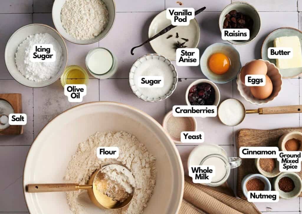 Various baking ingredients, including those for Air Fryer Hot Cross Buns&mdash;flour, sugar, yeast, eggs, butter, milk, spices, cranberries, raisins&mdash;are neatly arranged on a countertop with each item labeled. Utensils are also included.