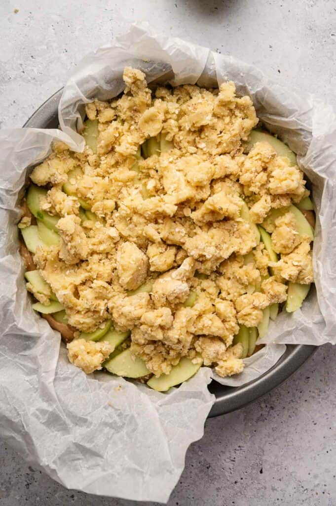 A round baking pan lined with parchment paper holds sliced green apples topped with an uneven layer of crumbly dough, ready to bake into a classic Irish Apple Cake.