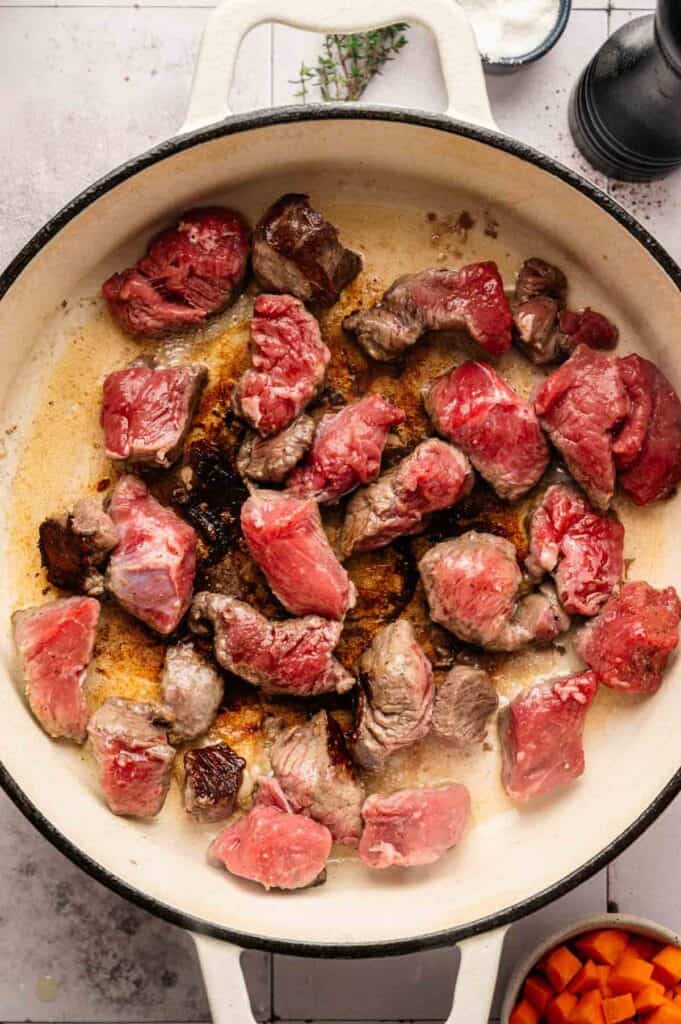 Cubed beef browning in a cast iron pot with thyme nearby