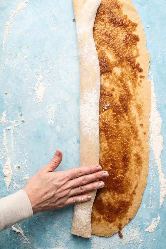 A hand rolls up dough spread with cinnamon and sugar filling on a floured blue surface, preparing a classic King Cake.
