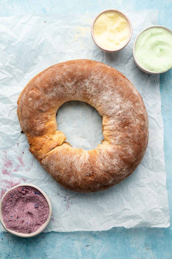 A round, golden-brown King Cake with a hole in the center sits on parchment paper next to three small bowls filled with colored sugar: purple, yellow, and green.