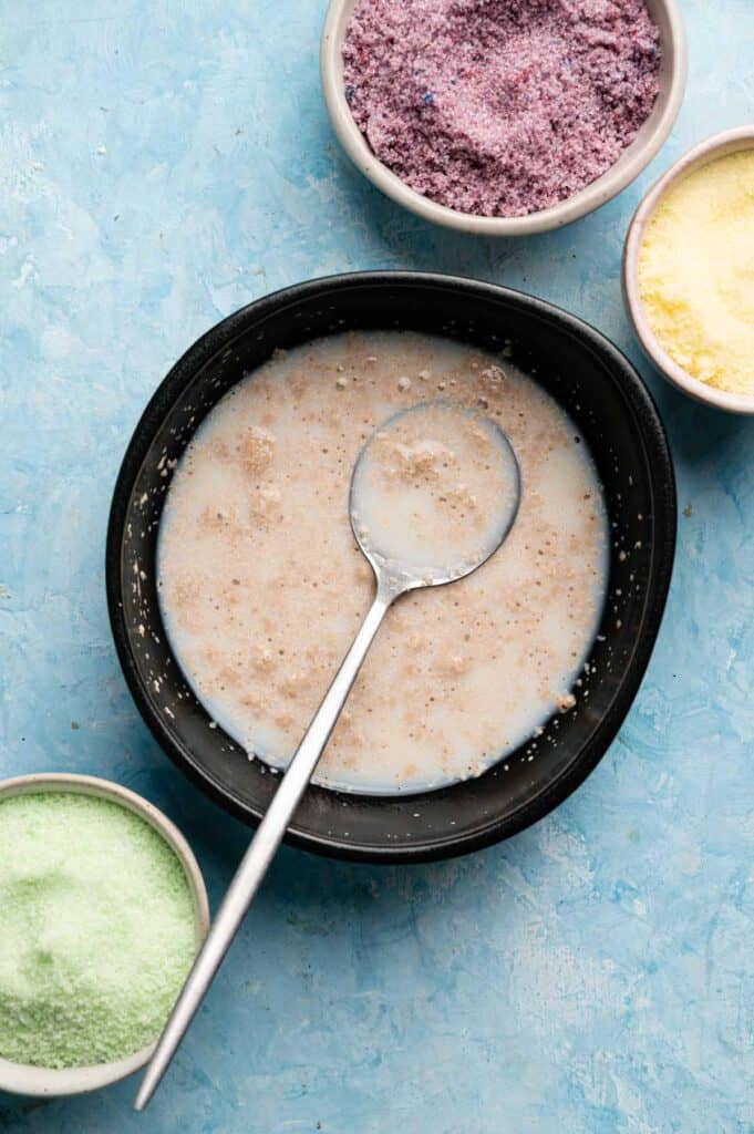 Foamy yeast mixture in a black bowl next to bowls of colored sanding sugar