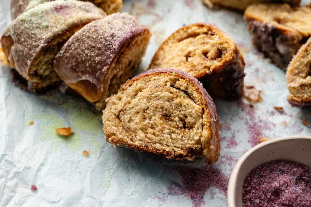 Sliced pieces of swirled King Cake bread with sugar topping rest on parchment paper, with colored sugar sprinkled nearby and a small bowl partially visible.