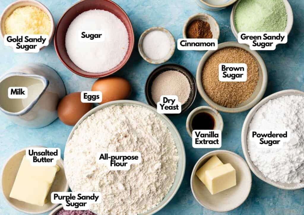 A variety of King Cake baking ingredients in bowls and dishes, including eggs, flour, butter, sugars, milk, dry yeast, cinnamon, and vanilla extract, arranged on a blue surface.