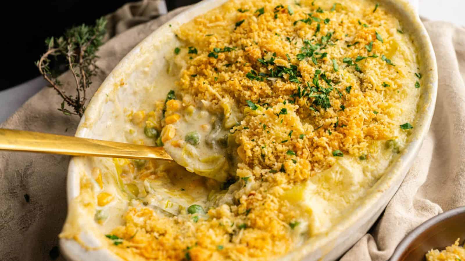 A casserole dish filled with creamy mixed vegetables topped with golden, crispy breadcrumbs and garnished with chopped herbs, partially served with a spoon.