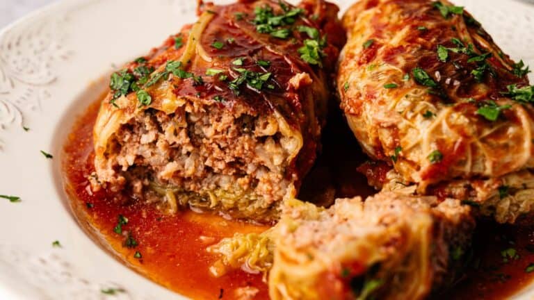 Two stuffed cabbage rolls topped with tomato sauce and chopped parsley, one cut open to show the ground meat and rice filling on a white plate.