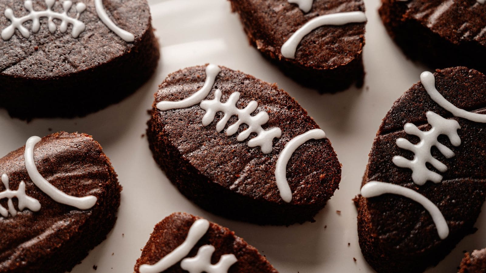 Oval-shaped brownies decorated with white icing to resemble footballs, arranged on a white surface.