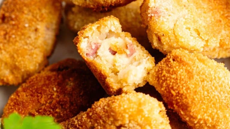 Close-up of several golden-brown breaded croquettes, with one broken open to reveal a creamy filling with small pieces of ham inside.