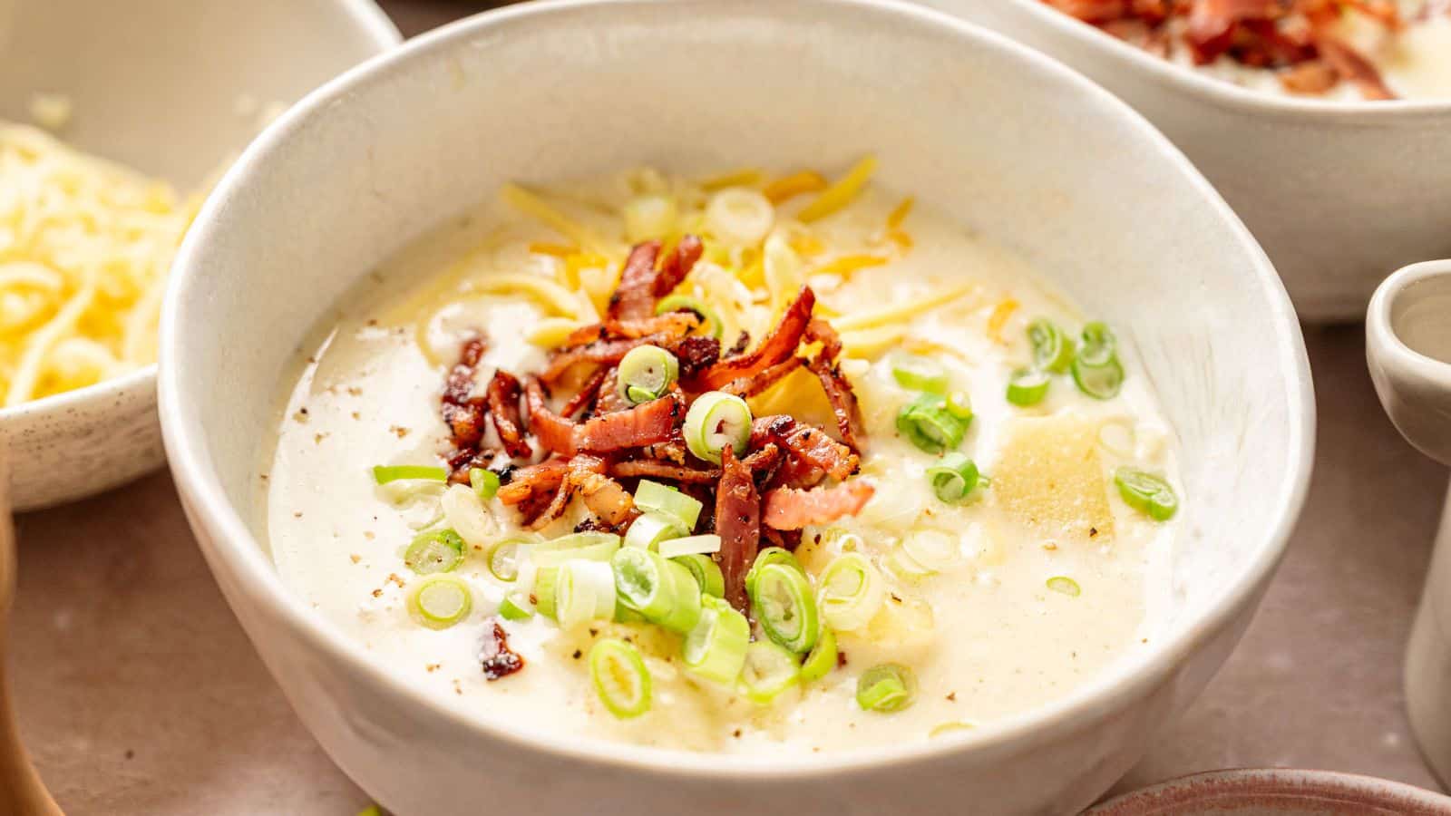A bowl of creamy soup topped with shredded cheese, chopped green onions, and crispy bacon pieces.