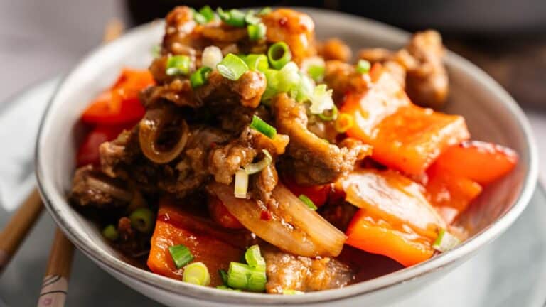 A bowl of stir-fried beef with red bell peppers, onions, and chopped green onions, served on a plate with chopsticks nearby.