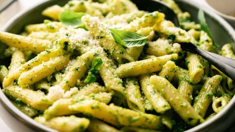 A bowl of penne pasta coated in green pesto sauce, topped with grated cheese and fresh basil leaves, served with a black spoon.
