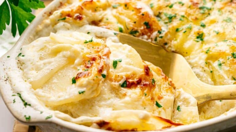 A close-up of creamy scalloped potatoes in a baking dish, garnished with chopped parsley, with a serving spoon lifting a portion.