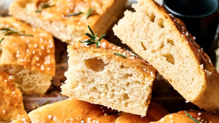Close-up of sliced focaccia bread topped with coarse salt and rosemary sprigs, showing its airy texture and golden crust.