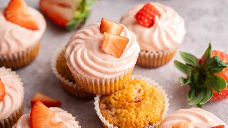 Cupcakes with pink frosting and sliced strawberries on top are arranged on a light surface, with whole and sliced strawberries scattered around.