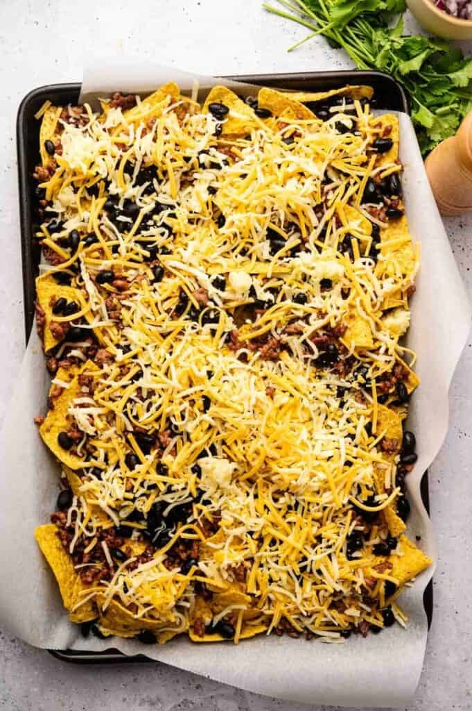A baking tray lined with parchment paper holds Sheet Pan Nachos topped with shredded cheese, black beans, and ground meat, ready to be baked.