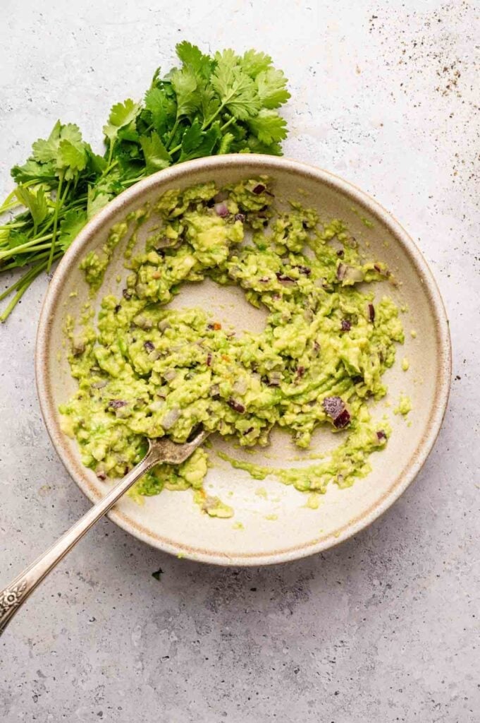 A bowl of freshly prepared guacamole with visible red onion pieces sits ready, perfect for pairing with sheet pan nachos, with a fork and cilantro garnished on the side.