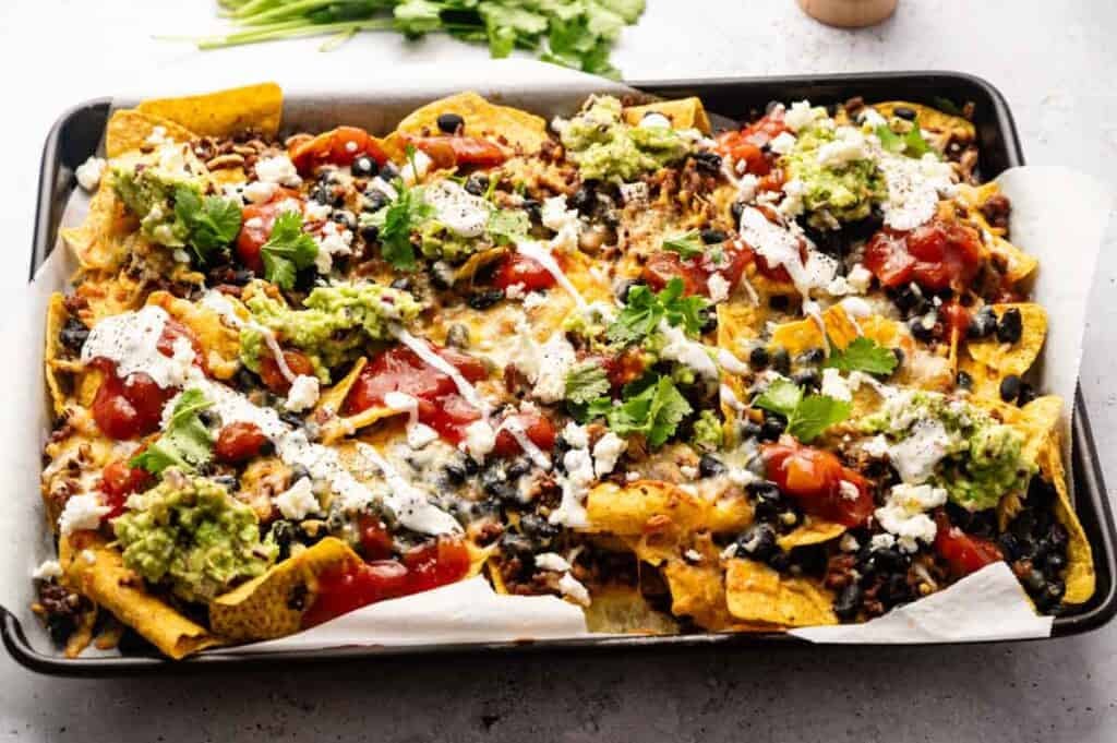 A tray of Sheet Pan Nachos topped with melted cheese, black beans, salsa, guacamole, sour cream, and fresh cilantro sits on parchment paper.